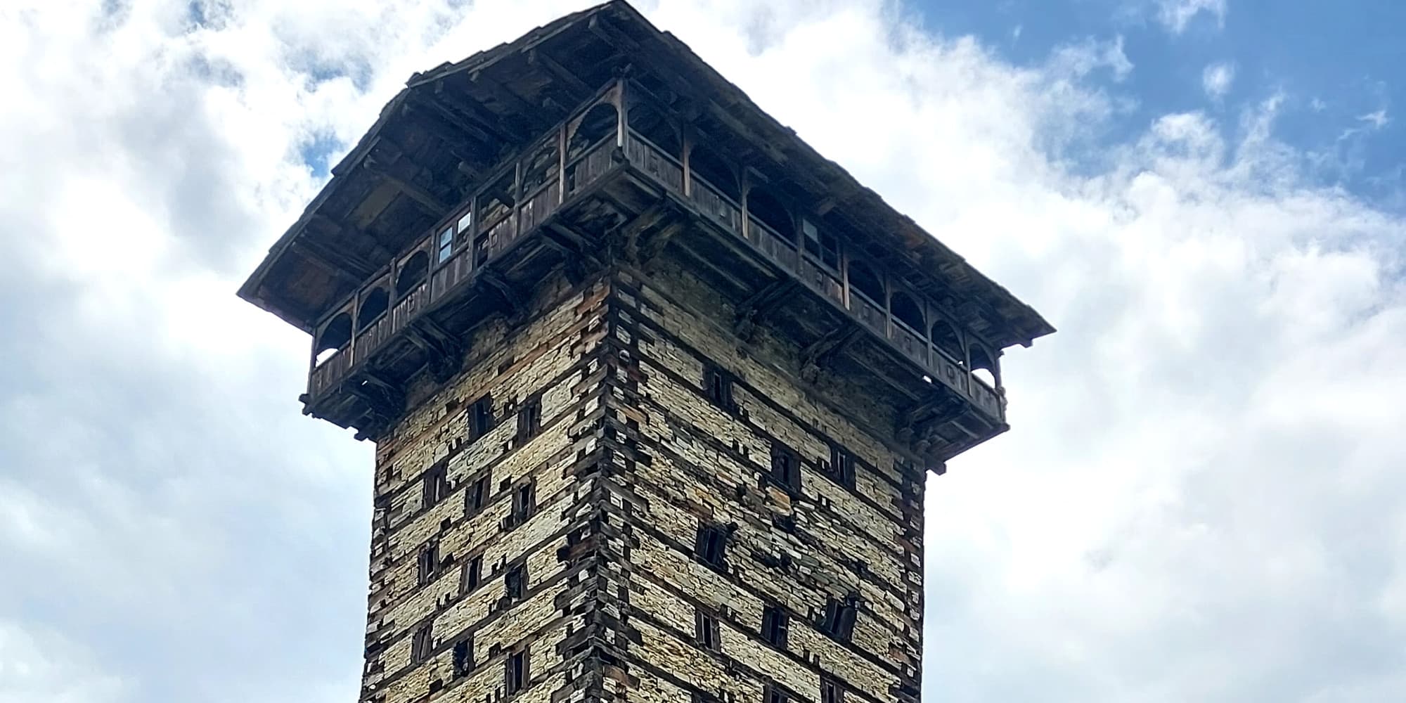 Upper stone and timber tiers of Chehni Kothi beneath a cloudy blue sky