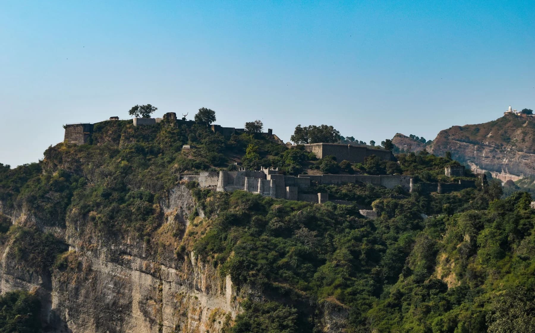 Kangra Fort's stone walls above a steep rocky hillside, with trees around the ruins