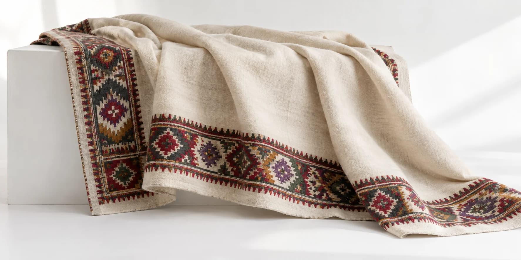 AI-generated illustration of a cream woollen wrap with colourful geometric borders draped on a white plinth
